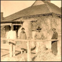 A sepia tone monochrome photo of four family members standing in their front lawn in front of a small house.