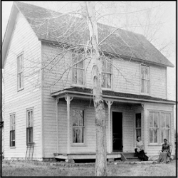 A black and white photo of a two story white house with a porch, and a baren tree in the front.